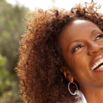 HPV vaccine: Close-up of a smiling Black woman with curly hair looking up towards the sky, wearing hoop earrings.
